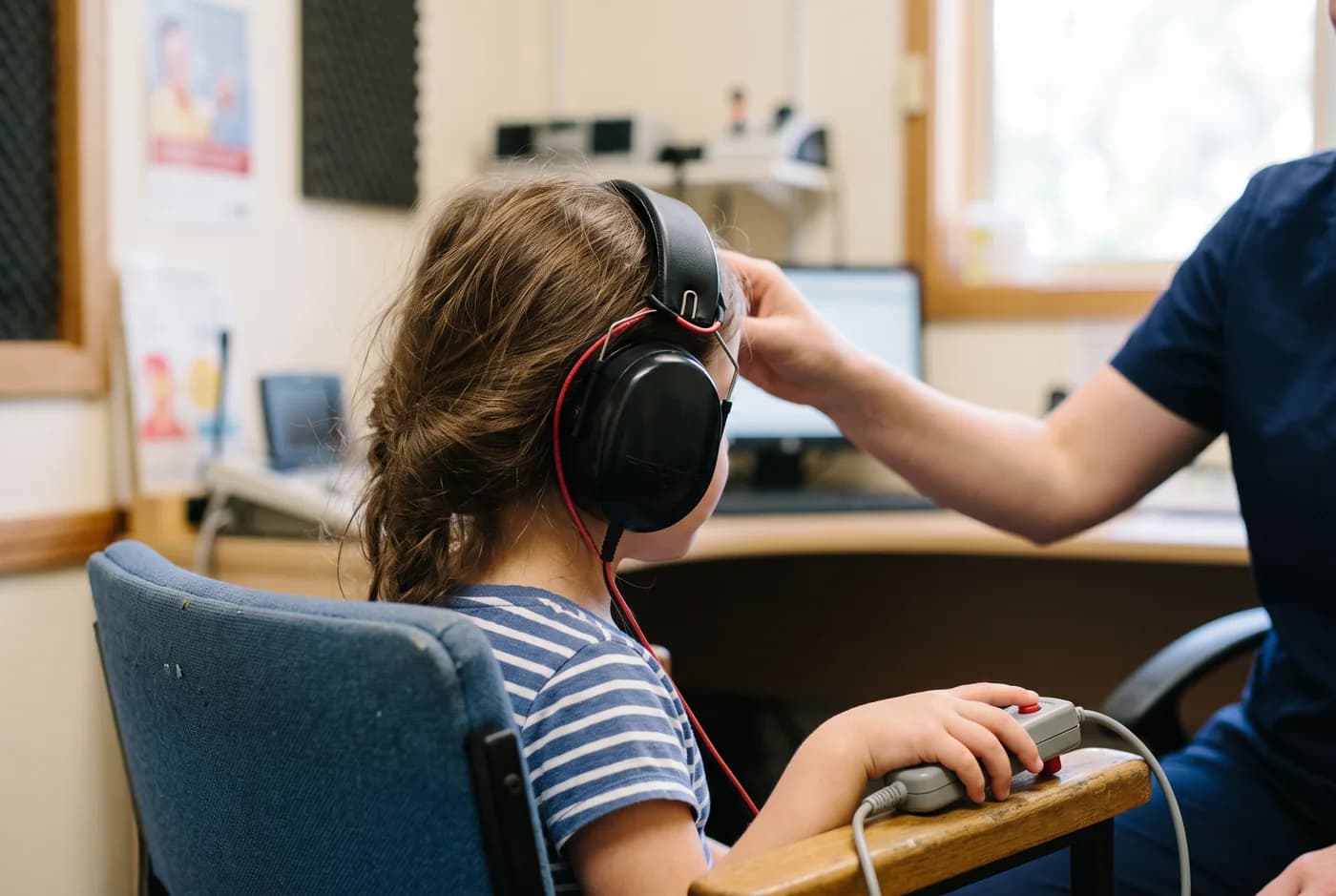 A child wearing test headphones and holding a response button while an audiologist adjusts the headband.